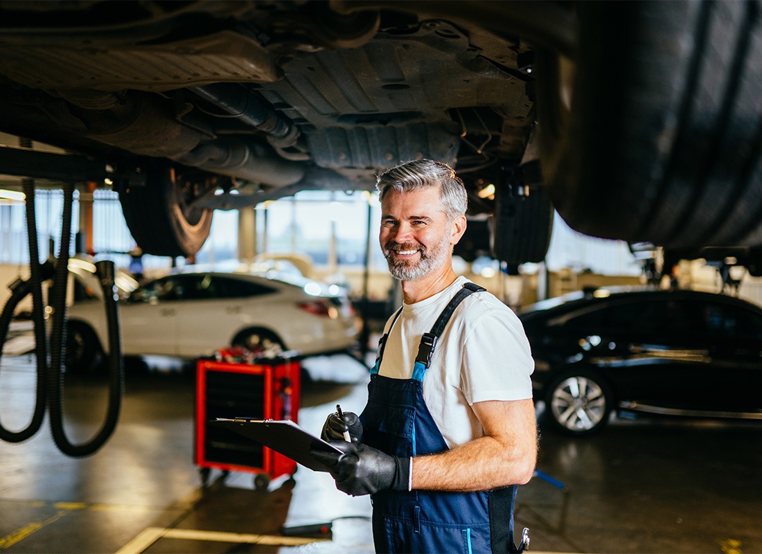Business Insurance - Portrait of a Cheerful Mature Business Owner Holding a Clipboard in his Hands as he Stands Underneath a Vehicle in his Garage Repair Shop